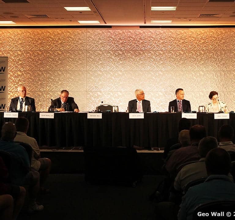 Panel of eight people seated behind a long table on stage, with one person standing and speaking at a podium labeled