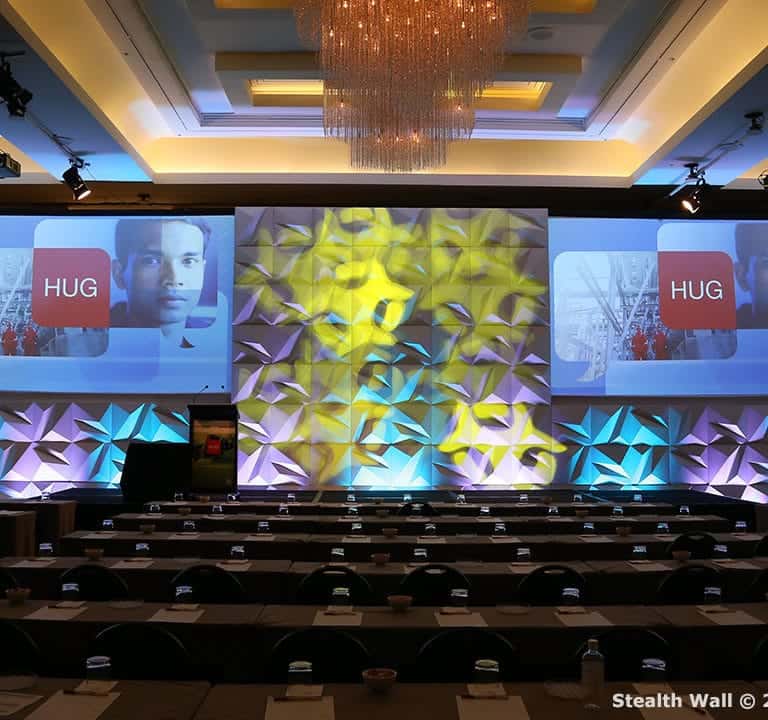 The conference room is set up with rows of tables and chairs facing a stage, featuring a large screen displaying a presentation. The backdrop, using Stealth Modular Backdrops, incorporates geometric patterns enhanced by blue and yellow lighting effects.