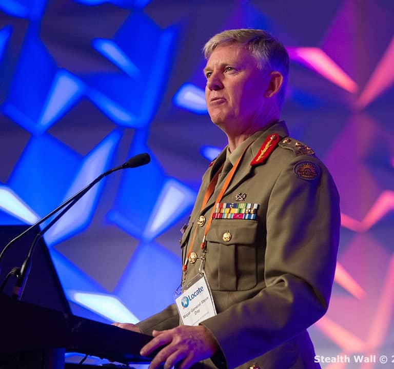 A military officer in uniform stands at a podium, speaking with a colorful geometric stage backdrop behind him, enhancing the event theming.