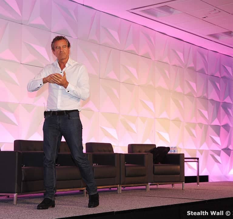 A man in a white shirt and jeans speaks on a stage with a geometric backdrop and chairs, perfectly capturing the event theming. There is a large screen displaying his image on one side.