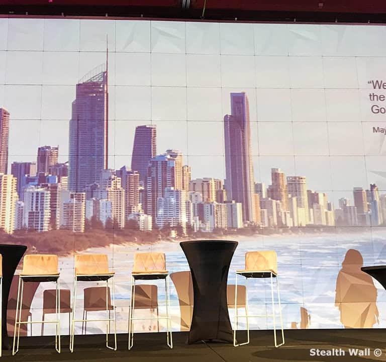 A man and woman stand on a stage with six empty chairs, framed by expertly designed stage backdrops. Behind them, a large image of a city skyline is projected. A quote by Mayor Tom Tate about investing in the future underscores the thoughtful event theming.