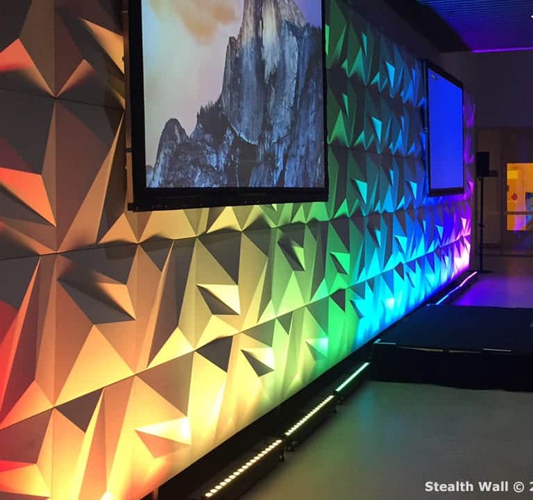 A geometric-patterned wall illuminated with gradient lighting in a range of colors, featuring two projection screens displaying an image, and a stage setup in front of it utilizes stealth modular backdrops for a seamless and dynamic visual experience.