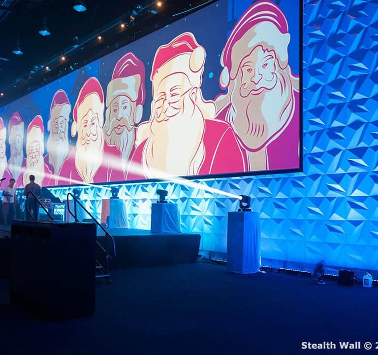 A stage decorated with a large screen displaying multiple Santa Claus images, blue geometric pattern walls, and Christmas trees with lights.© 2017 Stealth Modular Backdrops.