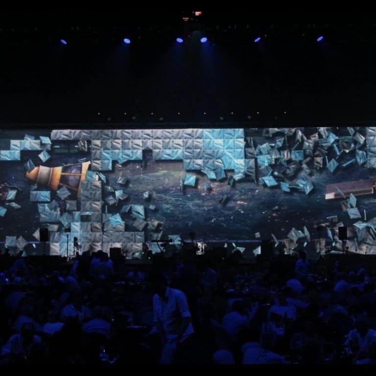 A large audience sits in a dark room facing an expansive, illuminated screen displaying a dynamic, fragmented visual with blue and geometric elements, enhanced by stealth modular backdrops.