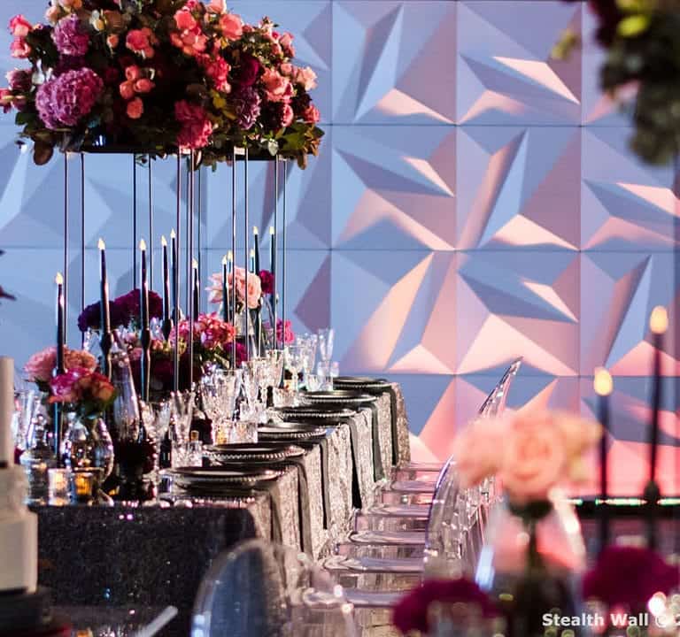 Elegant event setup with long banquet tables, floral centerpieces, clear chairs, and a tiered cake in the foreground against Stealth Modular Backdrops featuring a geometric-patterned wall.