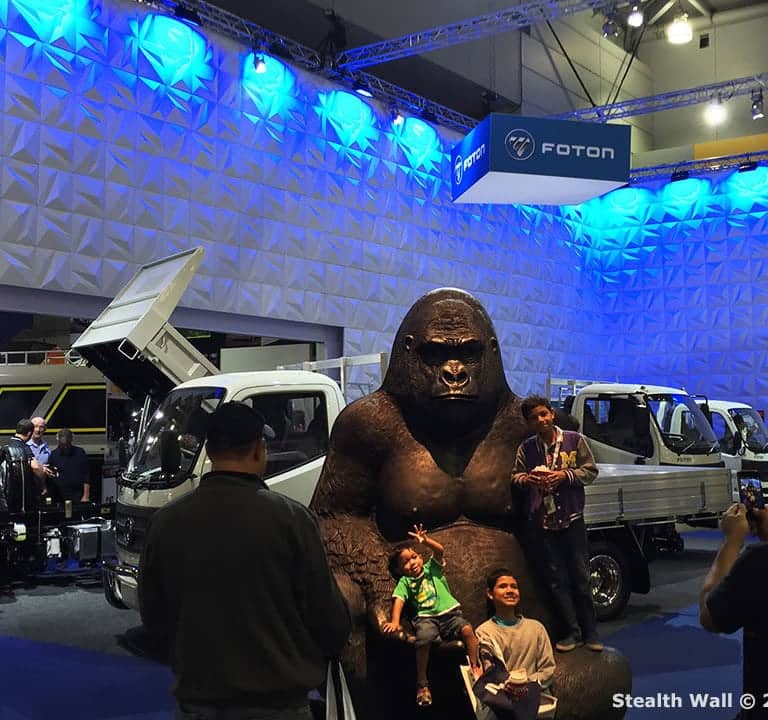 People are posing for a photo in front of a large gorilla statue at a booth featuring trucks, enhanced by Stealth Modular Backdrops, in an indoor exhibition space.