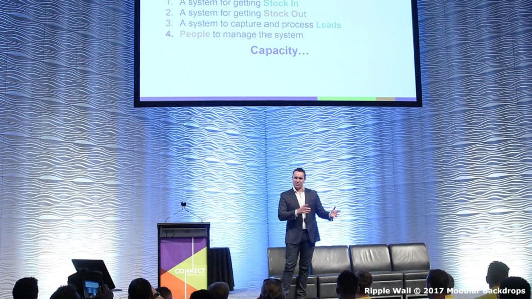 A man in a suit speaks on stage in front of a Ripple Wall backdrop and a large screen displaying text about system capacity. A podium with a laptop and a long sofa are also present on the stage.