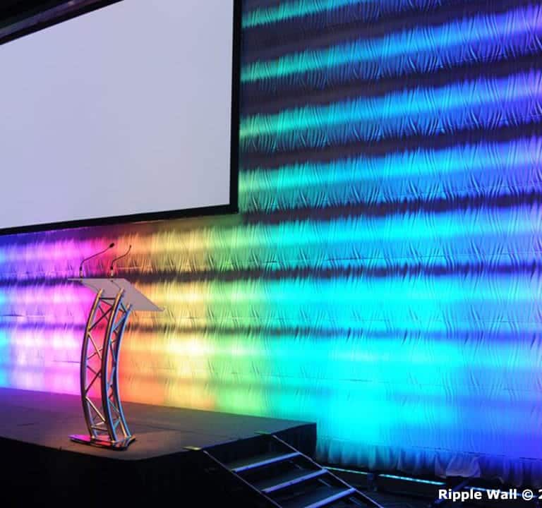 An empty stage with a podium stands in front of a large, colorful backdrop illuminated with rainbow lighting in a pattern. A projector screen hangs above the podium. The scene is enhanced by Ripple Wall Modular Backdrops that seamlessly blend into the vibrant ambiance.