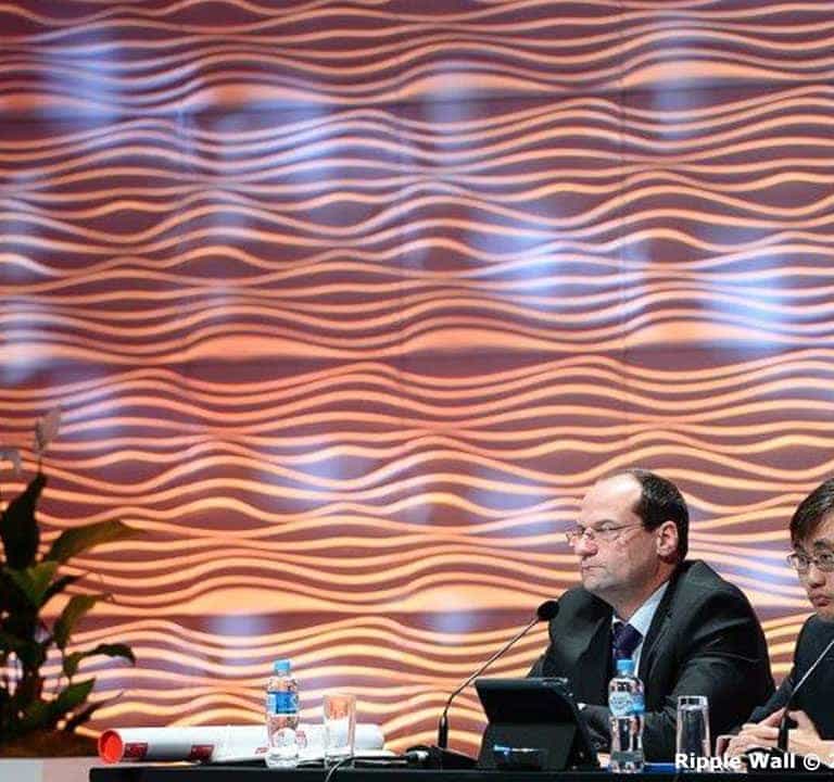 Two men sit at a conference table with microphones in front, framed by an elegant ripple wall. The textured wave patterns serve as a stunning backdrop, while laptops and a plant are also visible on the table, adding to the professional setting.