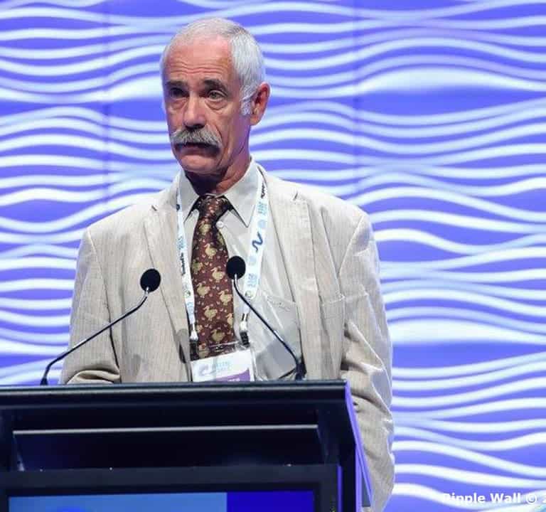 A man with a gray mustache wearing a light-colored suit speaks at a podium with Ripple Wall Modular Backdrops featuring a blue, wavy-patterned design.