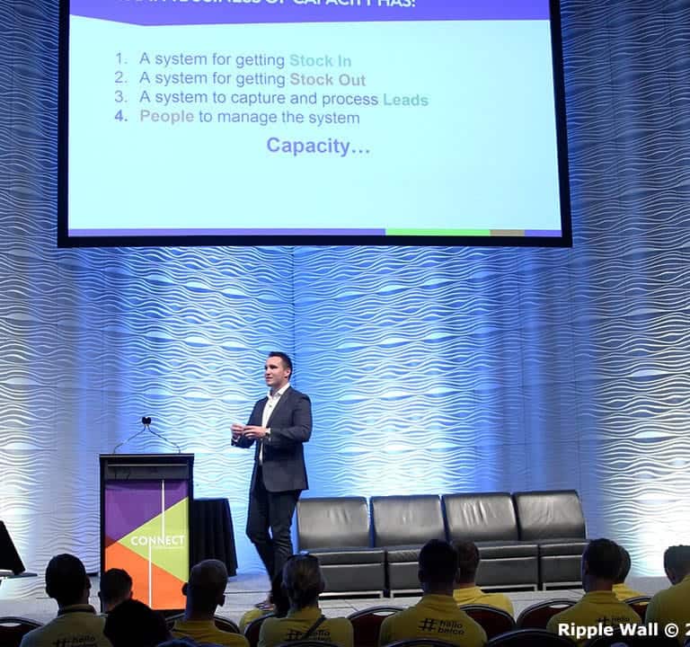 A man in a suit is presenting on stage with a projected slide behind him, framed by Ripple Wall modular backdrops. An audience in yellow shirts is seated facing the stage, attentively listening as the slide lists four systems related to capacity management.
