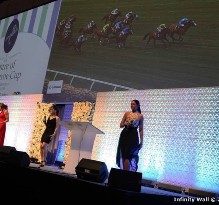 A stage event features four people at podiums in front of an illuminated infinity wall backdrop, with a large screen behind them displaying a horse race.