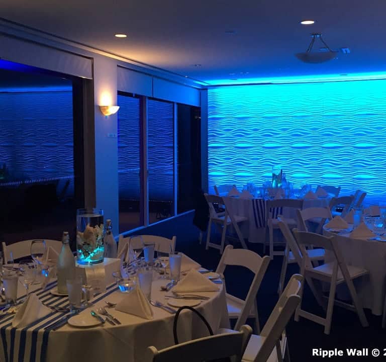 A dining room setup featuring round tables with white chairs, decorated with blue-lit ripple wall panels and Ripple Wall Modular Backdrops as stunning table centerpieces.