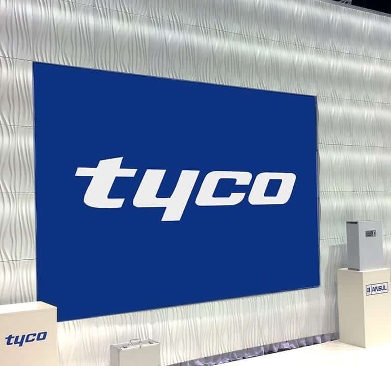 A large, blue Tyco logo displayed on a textured white wall with fire safety equipment and an Ansul sign in a trade show setting featuring Ripple Wall Modular Backdrops.