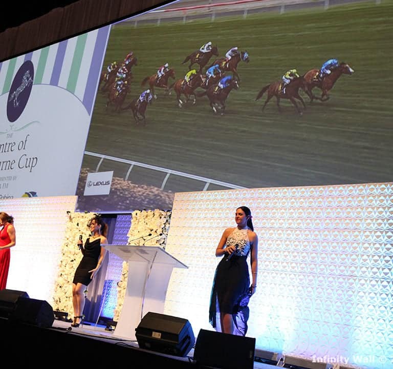A live band performs onstage while a large screen shows a horse race. Four performers, including one at a podium, are dressed in formal attire. The backdrop displays 