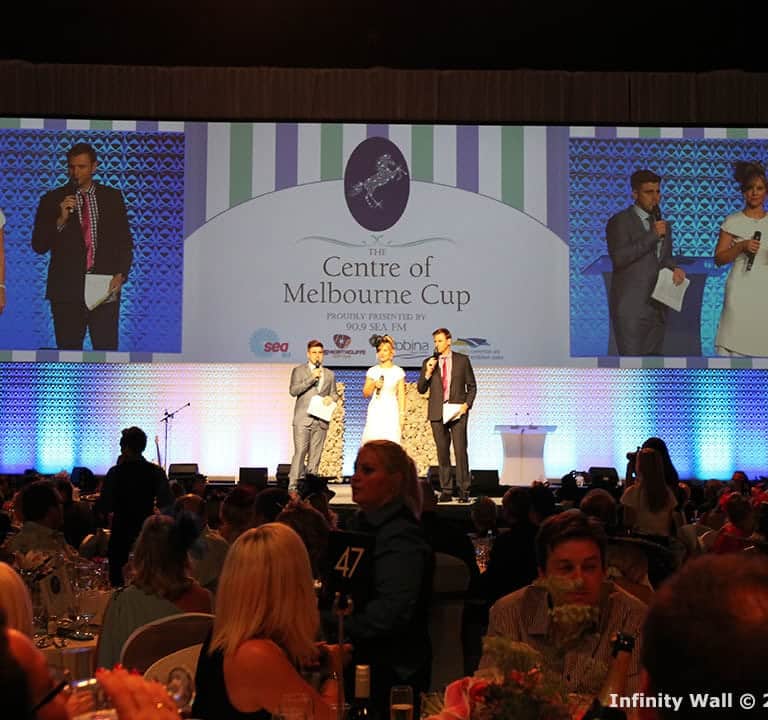 A stage presentation at the Centre of Melbourne Cup with four speakers holding microphones. Two large screens display the stage activity. Attendees are seated at tables in the foreground.