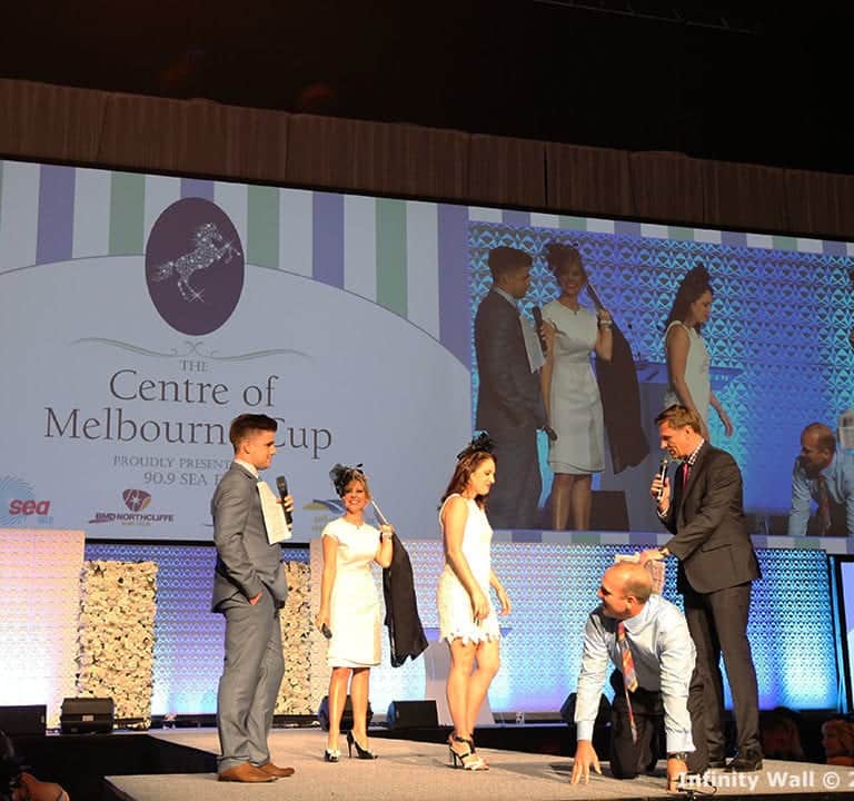 People are standing on an indoor stage under a projected display reading 