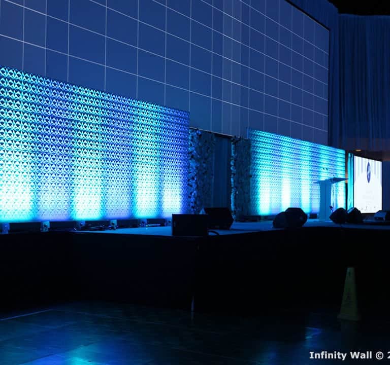 A stage with a large, illuminated backdrop featuring blue geometric patterns and a grid overlay. The stage is set up for an event with screens and lighting.