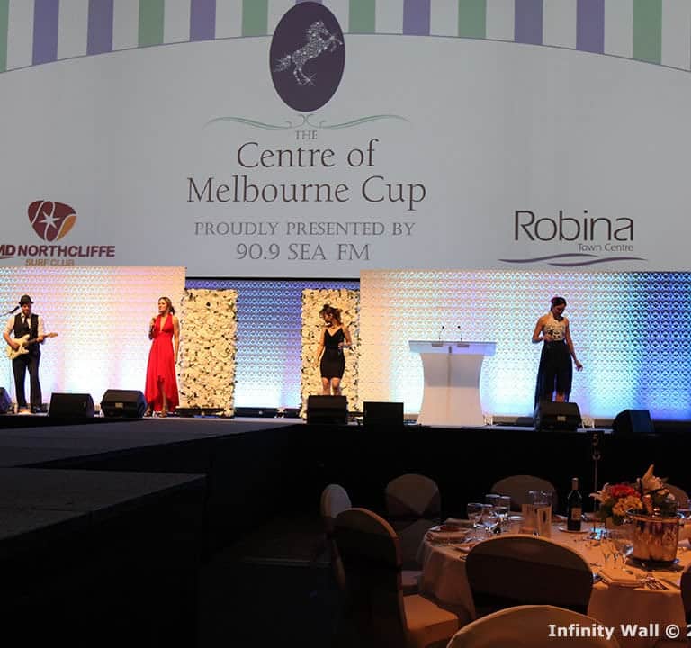 People on stage performing at The Centre of Melbourne Cup event, with a decorated background and round tables arranged in the foreground.