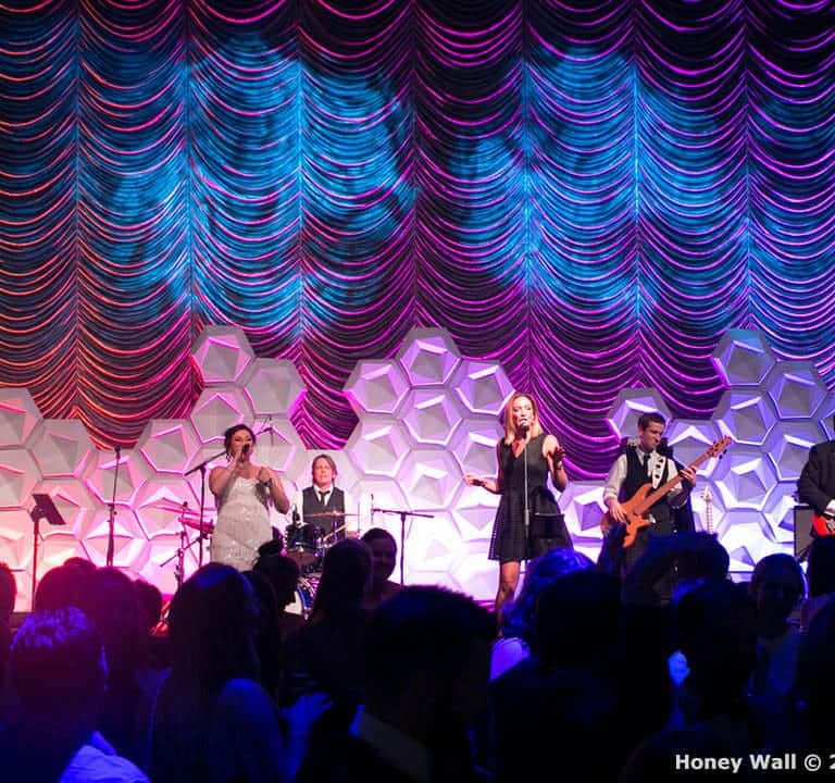 A band performs onstage under colorful lights. The backdrop features a geometric pattern. The audience stands in the foreground.