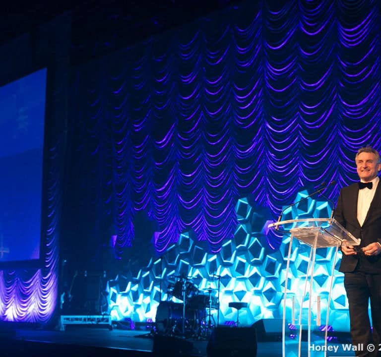 A person in a tuxedo speaks at a clear podium on a stage with a purple and blue draped backdrop and geometric decorations. Musical instruments are visible in the background.