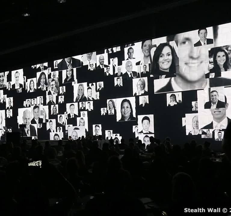 A large event with an audience seated in a dark room features a massive video wall displaying numerous black-and-white headshots of people. The display is labeled