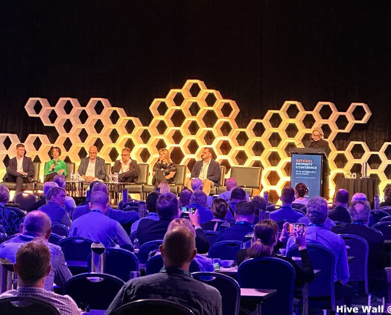 A panel of speakers sits on stage at a conference against a Hive Modular Panel backdrop, resembling a honeycomb. The audience is seated, and a presenter stands at a podium labeled