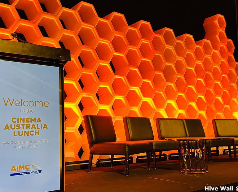 Stage setup with a Hive Modular Panel backdrop featuring a honeycomb pattern, podium, and chairs for the Cinema Australia Lunch. A screen displays event details.