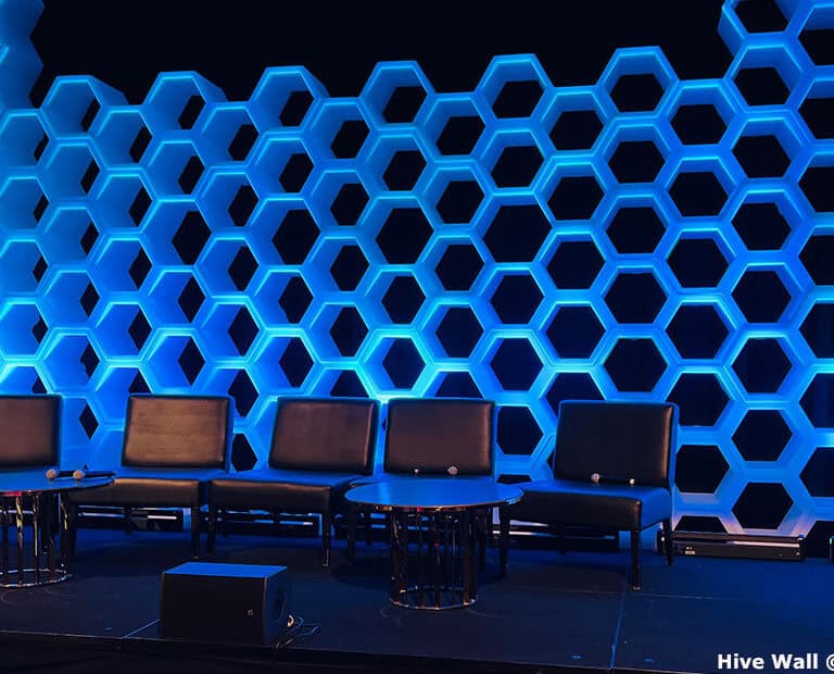 Stage setup with a hive modular panel as a honeycomb-patterned backdrop, multiple empty chairs, and two round tables, all beautifully lit in blue.