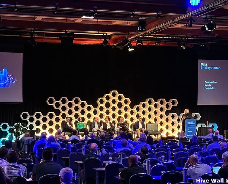 A conference stage with a Hive Modular Panel backdrop features panelists discussing data. Two large screens display a presentation slide, while attendees are seated and observing.