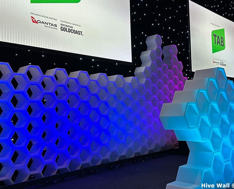 Illuminated hexagonal wall with blue and purple lighting in front of black backdrop featuring star-like lights and screens with logos.