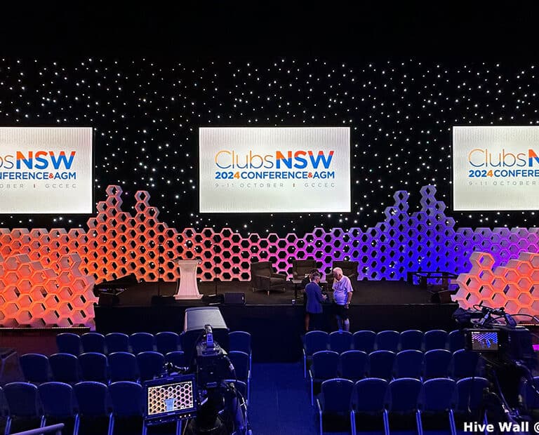Stage setup for the ClubsNSW 2024 Conference AGM features a hexagonal-patterned backdrop using Hive Modular Panels, complemented by three large screens and a spacious seating area.