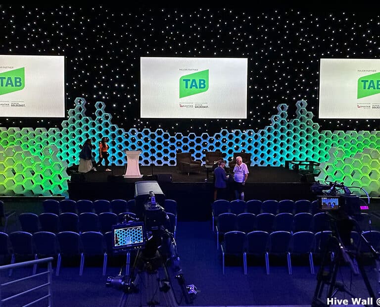 The conference stage setup features a Hive Modular Panel with a honeycomb-patterned backdrop, complemented by three screens showcasing