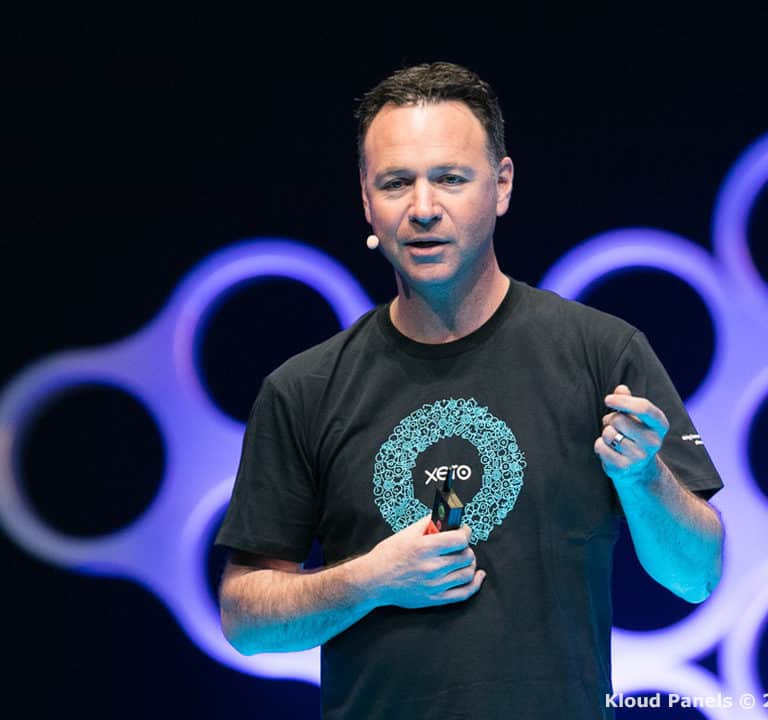 A man in a black t-shirt speaks into a headset microphone on a stage with a blue and purple geometric background. He holds a small object in his hand.