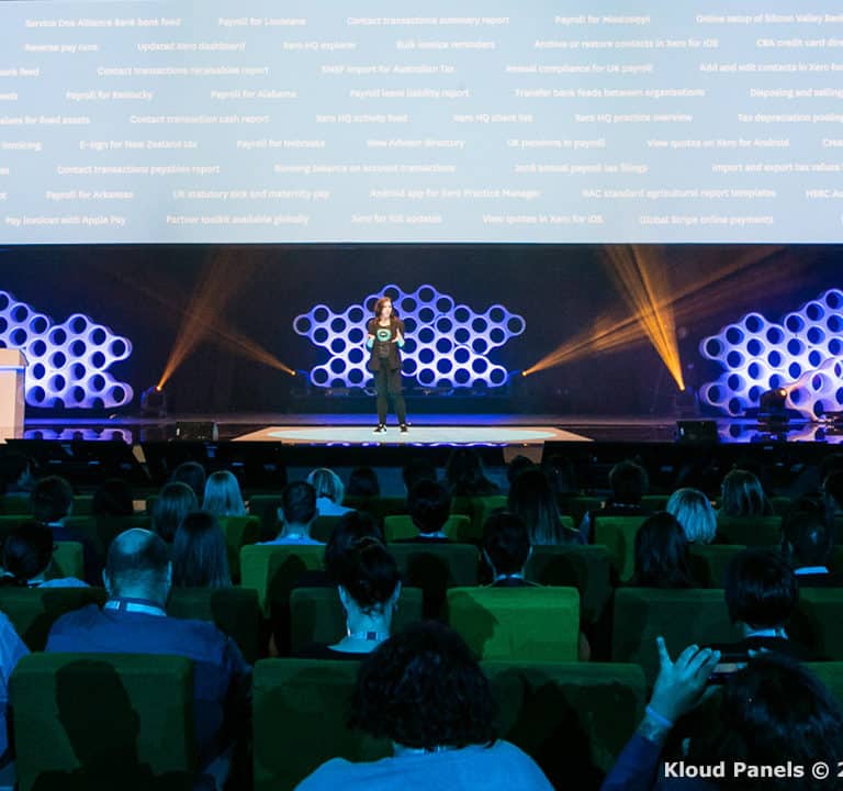A speaker on stage addresses a seated audience in an auditorium with a backdrop featuring honeycomb patterns and projected text. A podium stands to the left.
