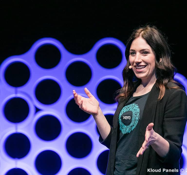 A woman wearing a blazer and a t-shirt is speaking on stage with circular design panels in the background.