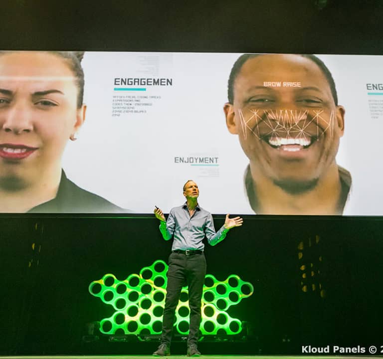 A speaker stands on stage in front of a large screen displaying images of people's faces with labeled emotions and facial recognition data.