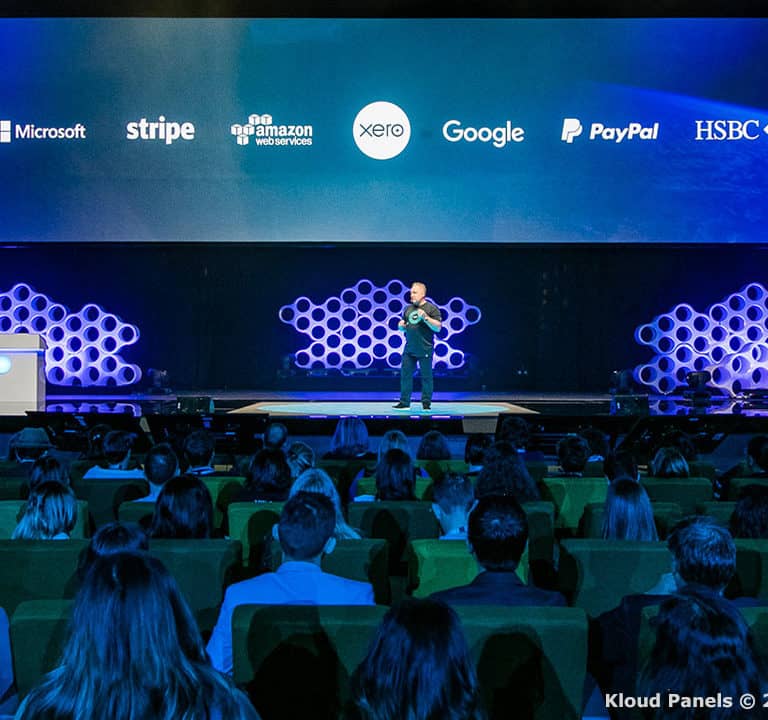 A speaker presents on stage at a conference with logos of major tech companies displayed on a screen behind them. The audience is seated facing the stage.