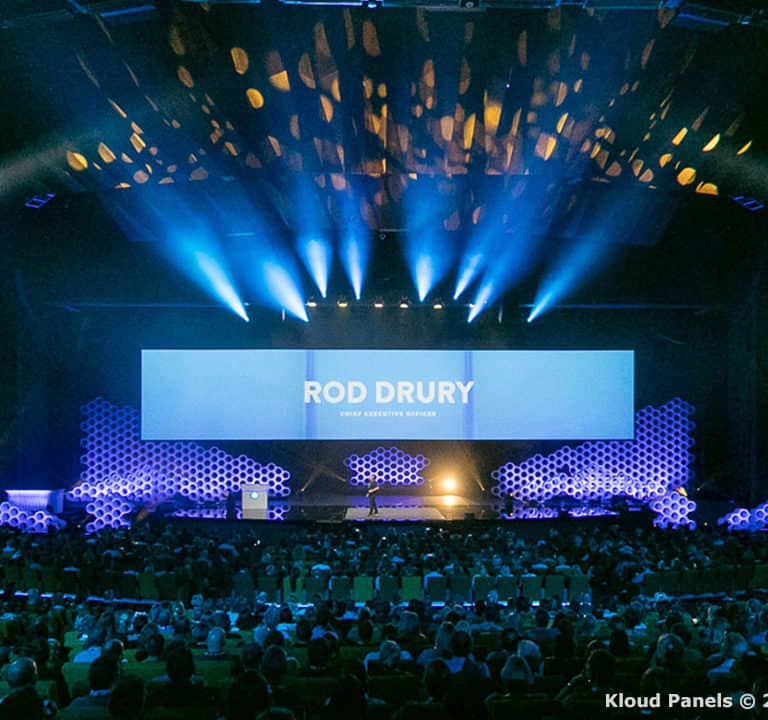 A stage with a large audience, blue lighting, and a screen displaying 