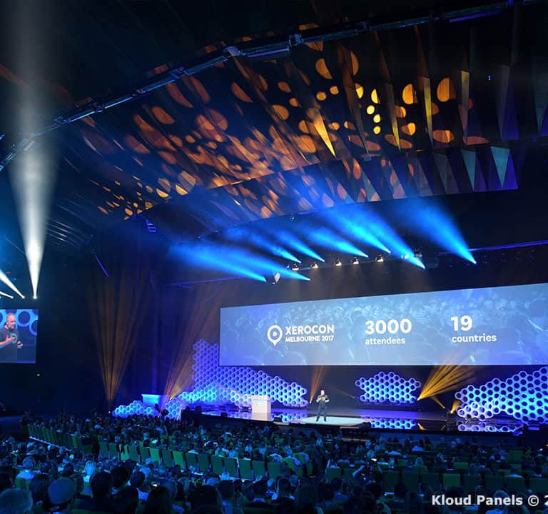 A large conference hall filled with an audience. A stage features a speaker and a large screen displaying 