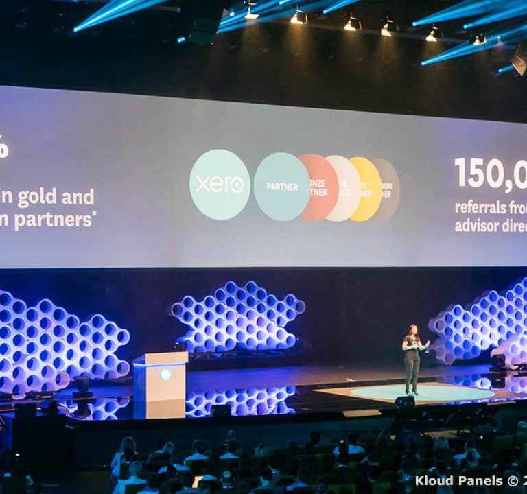 A person is speaking on stage in front of a large screen displaying business statistics and partner logos. The screen reads 