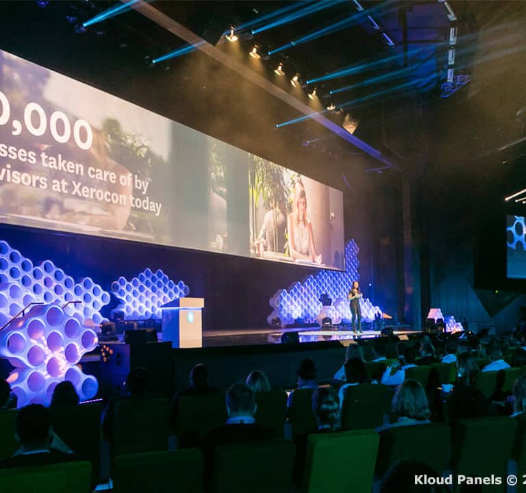 A conference room filled with attendees listening to a speaker on stage. A large screen displays 