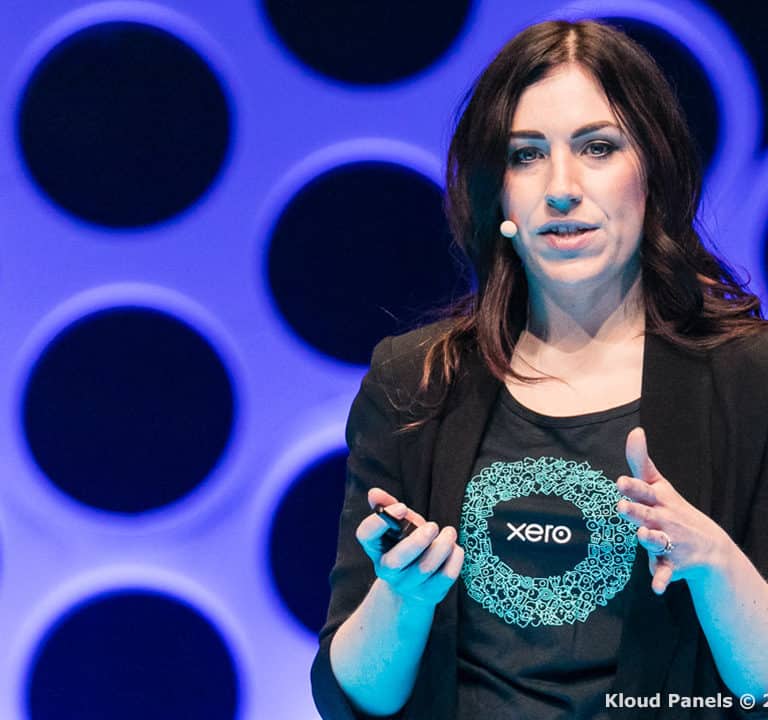 A person with a microphone headset speaks while gesturing and holding a device in one hand. The background features circular patterns and soft blue lighting.
