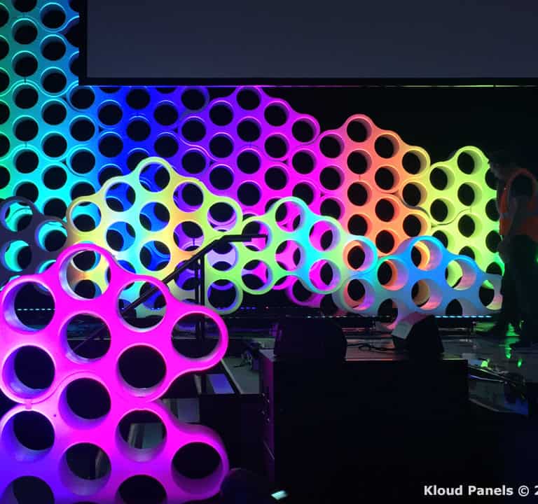 A vibrant stage with multicolored hexagonal panels illuminated in various hues. A person stands behind the panels, partially obscured by the light effects.