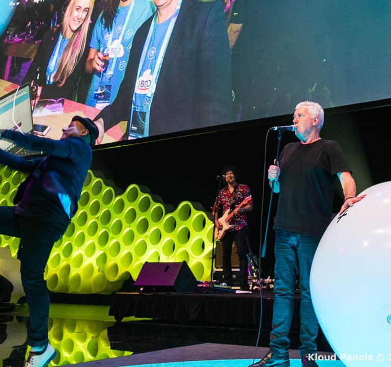 People on a stage with large white balloon props and a screen displaying an image of them. A person kicks one of the balloons while another stands near a microphone. A backdrop with hexagonal patterns is visible.