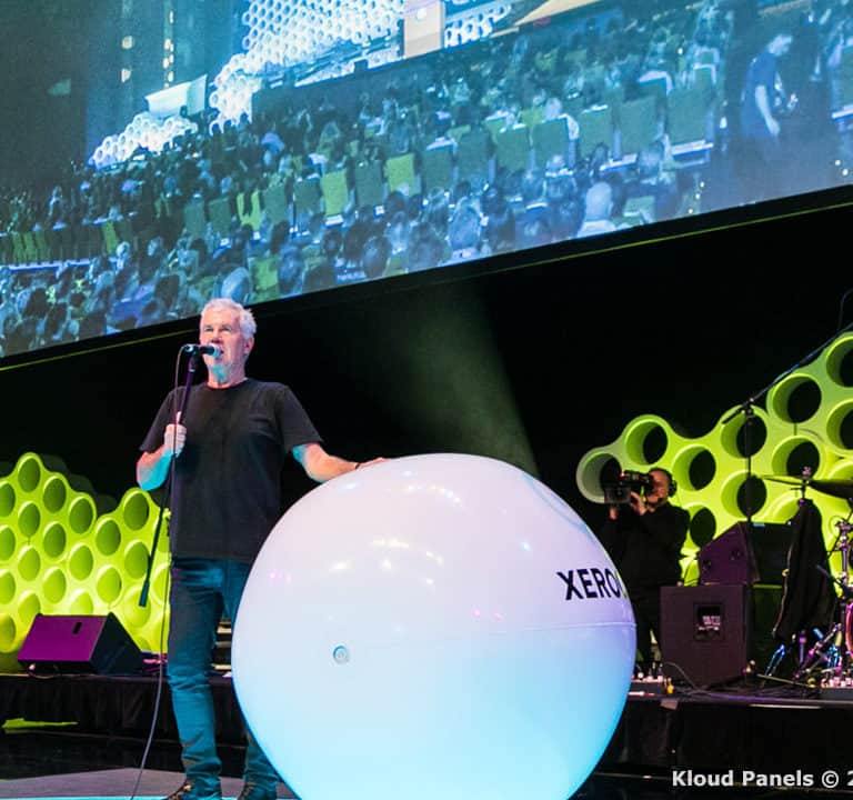 A man speaks into a microphone on stage next to a large white sphere. A live band plays nearby. The backdrop features a green hexagonal pattern with a large screen above displaying the stage scene.