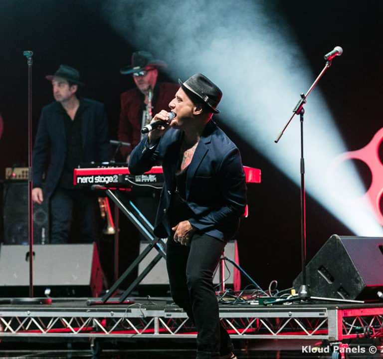 A band performs on stage with a lead singer in the foreground wearing a hat and suit, and musicians in the background playing instruments. The stage has red and white lighting and circular patterns.