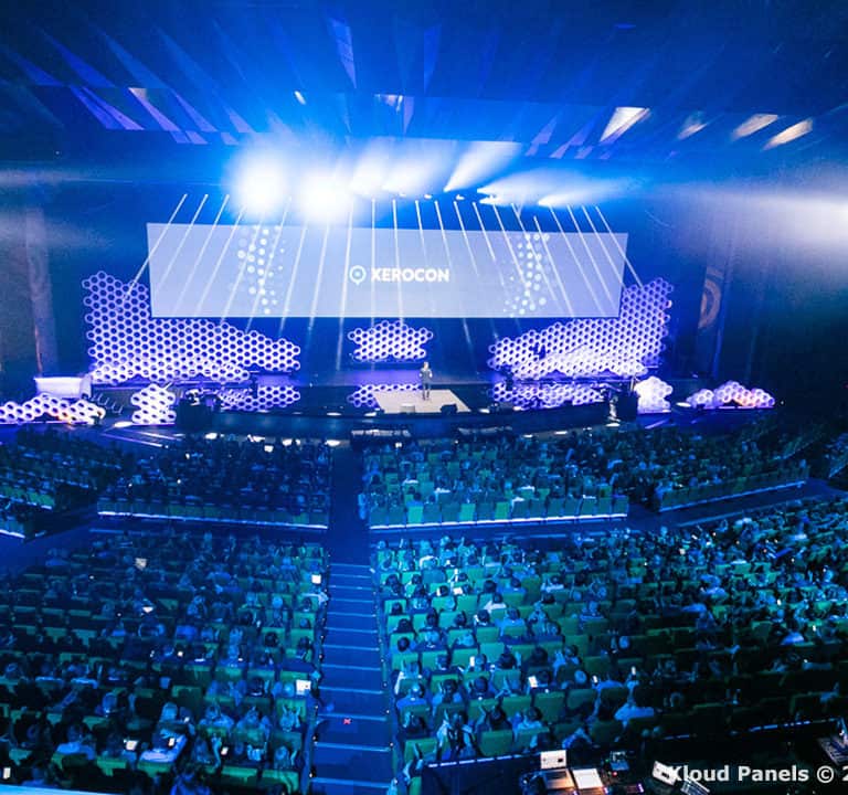 Wide-angle view of a conference hall filled with attendees, featuring a large stage with multiple screens displaying the word 