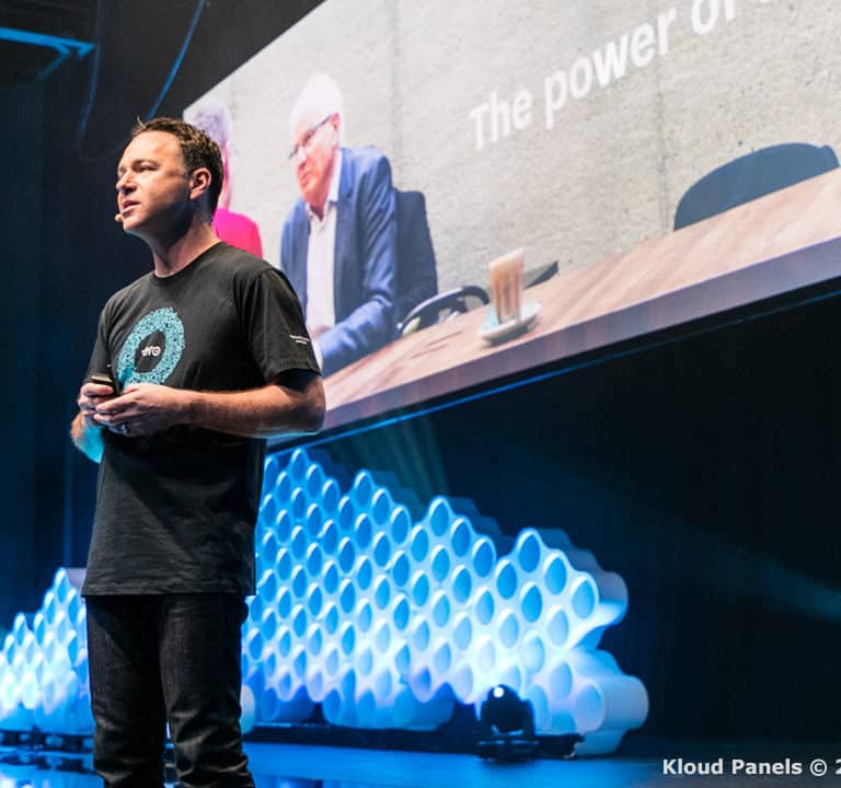 A man wearing a black T-shirt speaks on stage in front of a large screen with hexagonal light panels in the background.