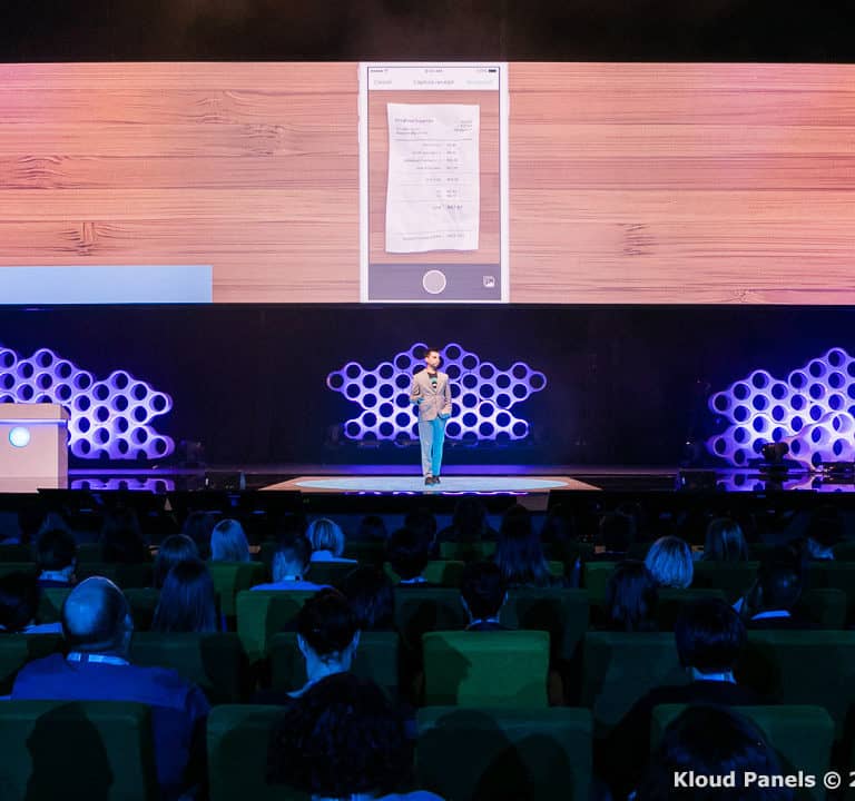 A person stands on stage presenting to an audience with a large screen behind displaying a document. The background features a honeycomb pattern.